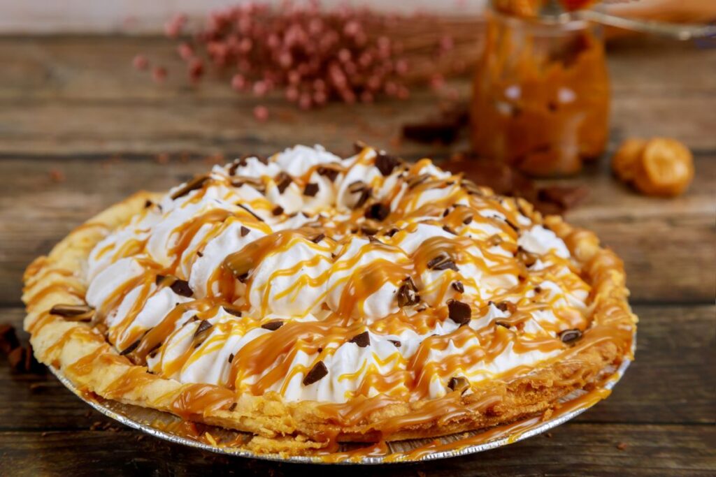 A homemade butterscotch pie topped with whipped cream, caramel drizzle, and chocolate shavings on a rustic wooden table.