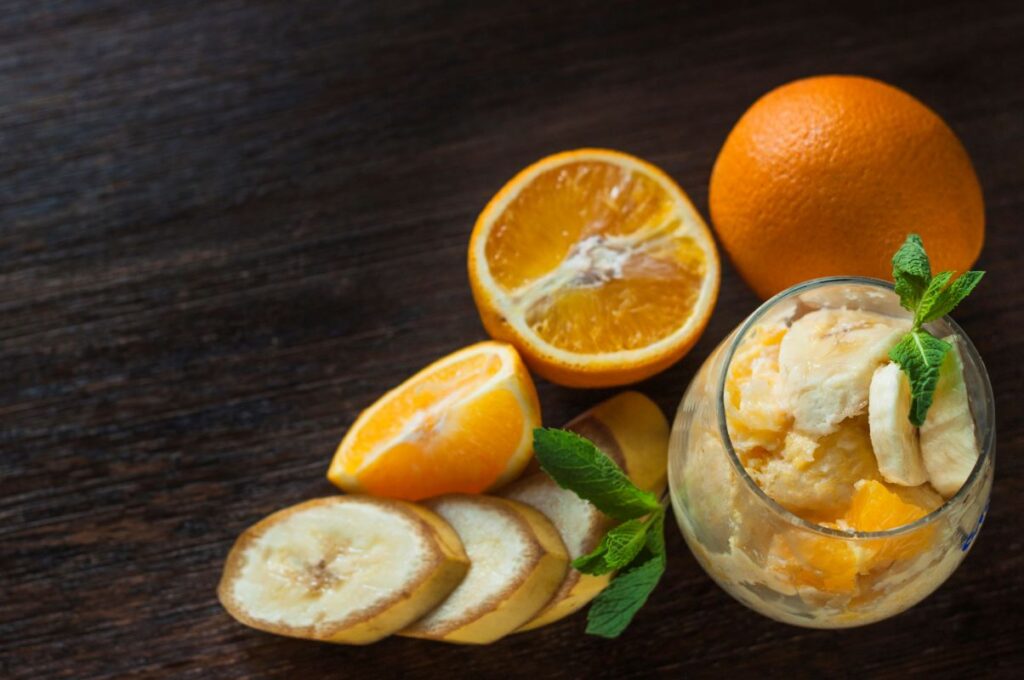 Overhead view of fresh oranges, banana slices, and a glass of orange creamsicle moonshine garnished with mint leaves on a dark wooden table.