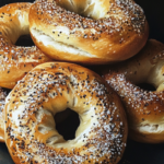 A close-up of a stack of golden-brown salt bagels with a sprinkle of coarse salt and sesame seeds on a dark background.